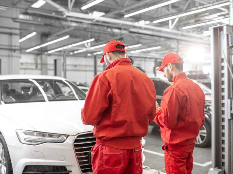 Two auto repair technicians in red uniforms working on a white car in a well-lit garage setting.