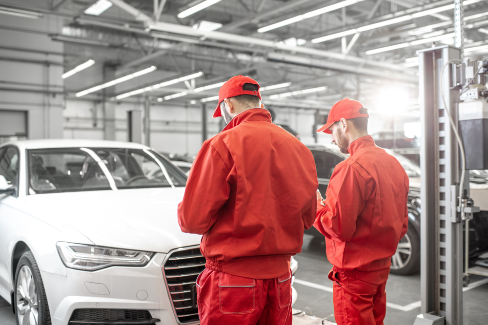 Two auto repair technicians in red uniforms working on a white car in a well-lit garage setting.