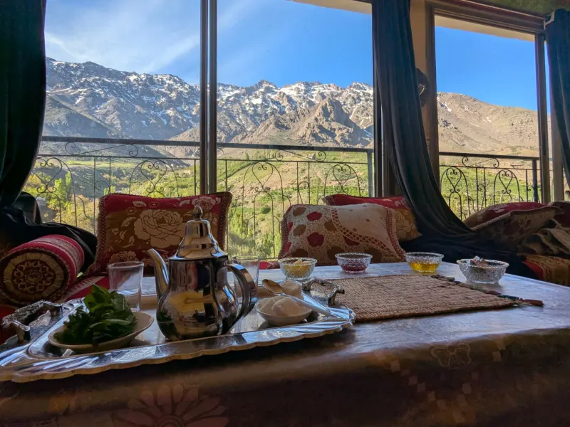 A cozy Moroccan tea setup with a stunning view of snow-capped mountains and vibrant green landscapes through large windows.