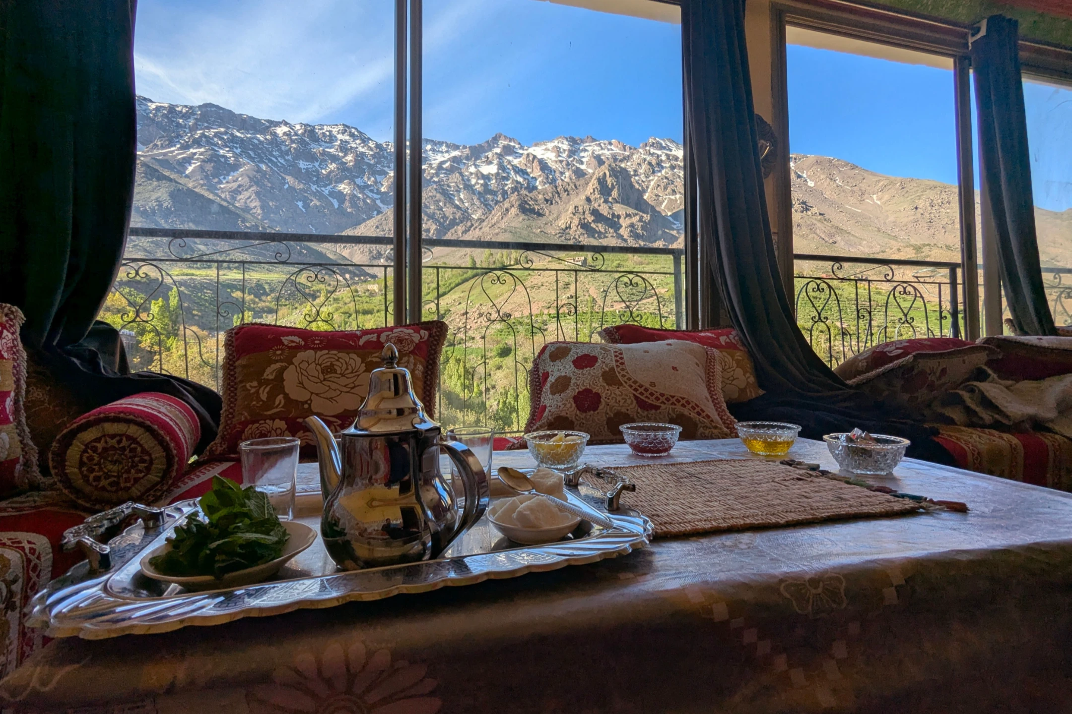 A cozy Moroccan tea setup with a stunning view of snow-capped mountains and vibrant green landscapes through large windows.