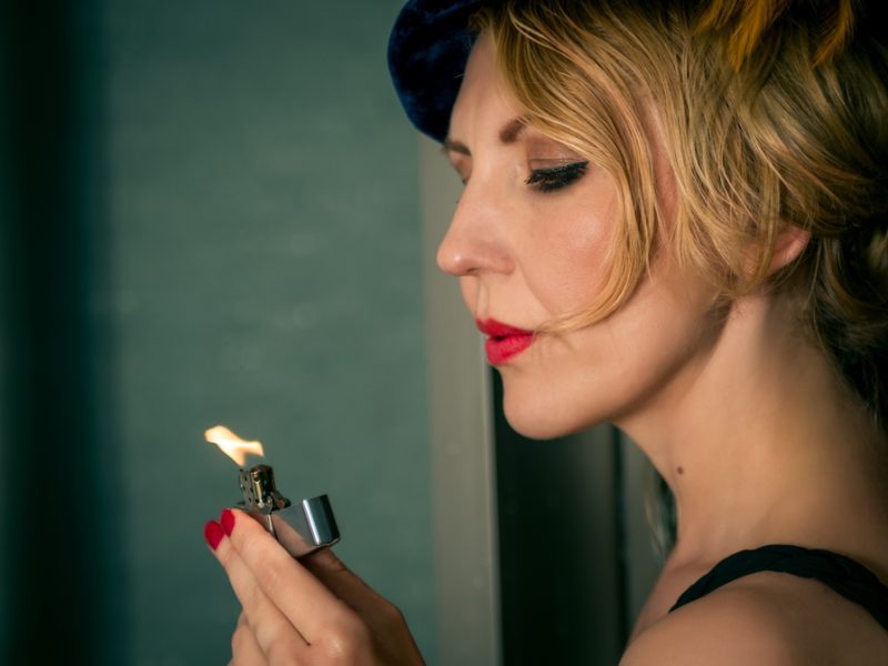A woman with red nails lights a silver lighter while wearing a black dress and a stylish blue hat.
