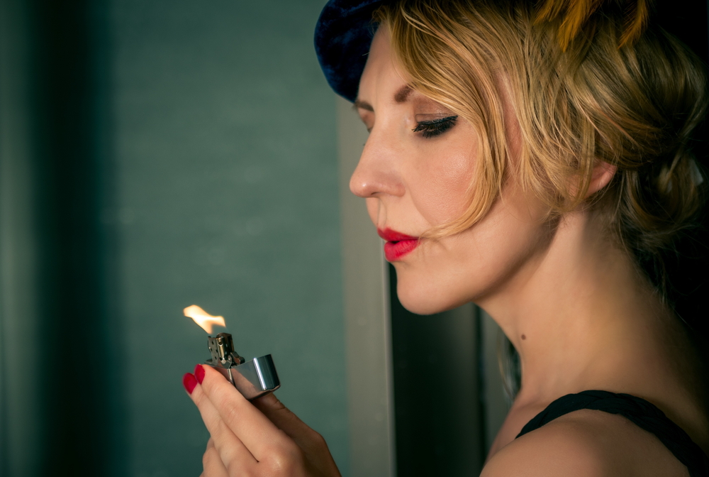 A woman with red nails lights a silver lighter while wearing a black dress and a stylish blue hat.