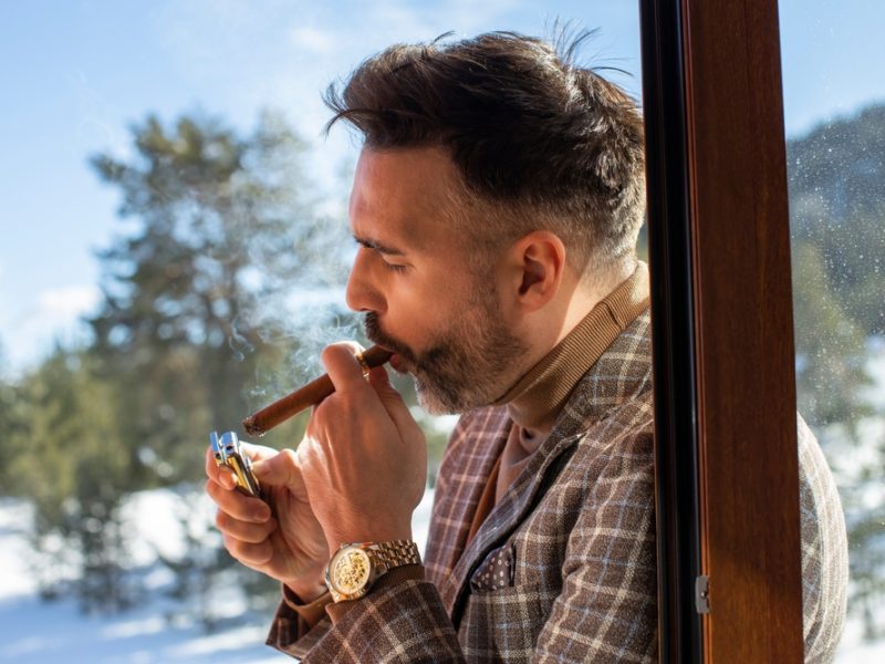 A man is lighting a cigar near a window, with snowy trees visible outside in the background.