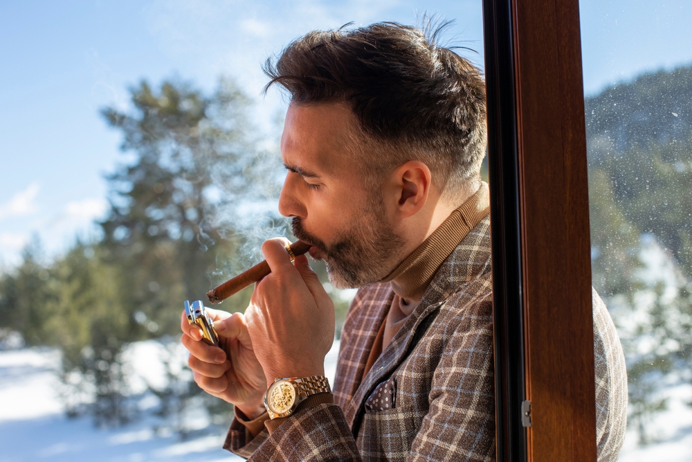A man is lighting a cigar near a window, with snowy trees visible outside in the background.
