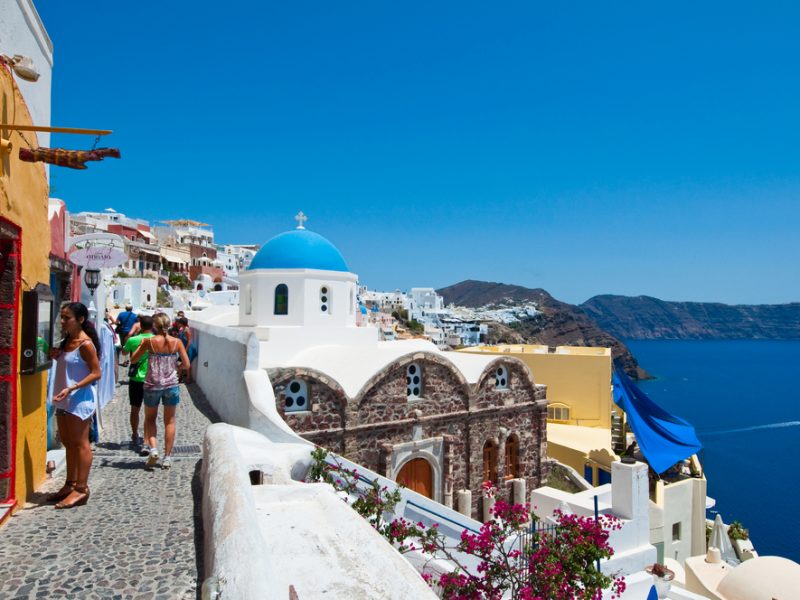 Crowded cobblestone street in Santorini with people exploring, colorful buildings, and a view of the caldera under a clear blue sky.