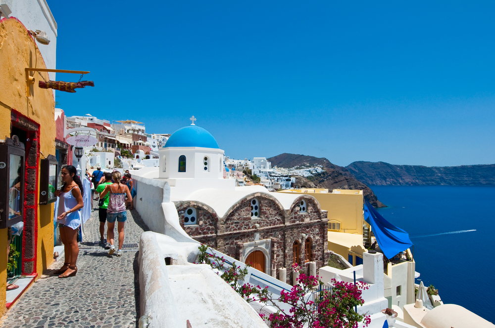 Crowded cobblestone street in Santorini with people exploring, colorful buildings, and a view of the caldera under a clear blue sky.