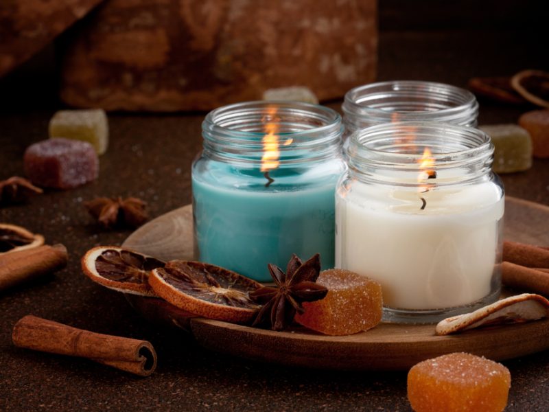 Three scented candles in glass jars, surrounded by dried orange slices, cinnamon sticks, and colorful candy on a wooden plate.