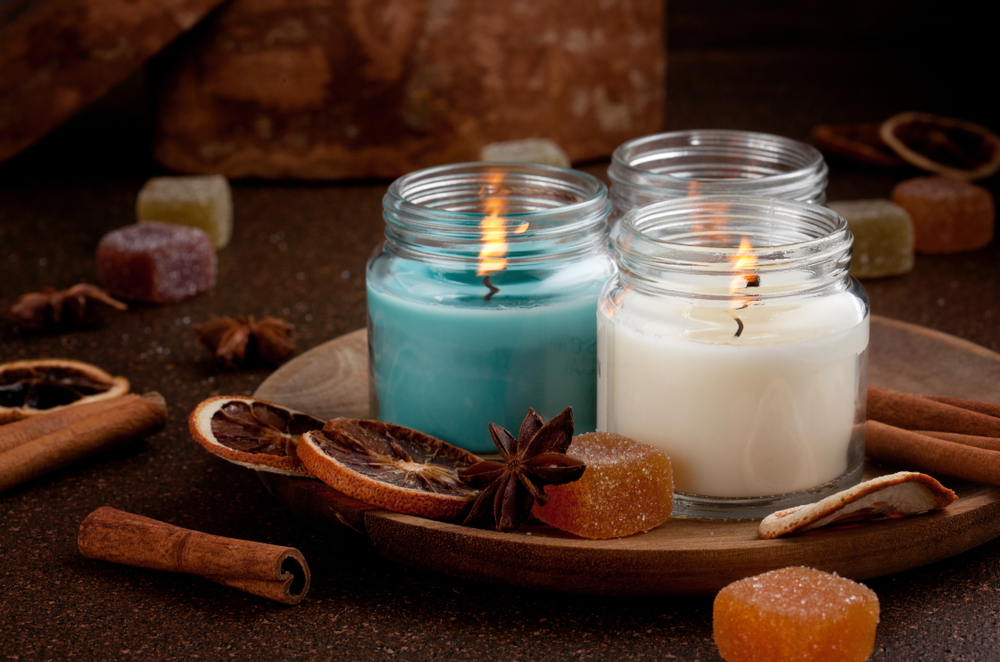 Three scented candles in glass jars, surrounded by dried orange slices, cinnamon sticks, and colorful candy on a wooden plate.