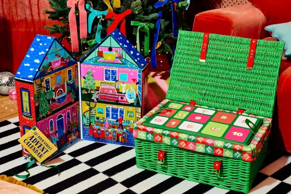 Colorful advent calendars designed by Lush, featuring a festive house and a green wicker basket, on a striped rug.