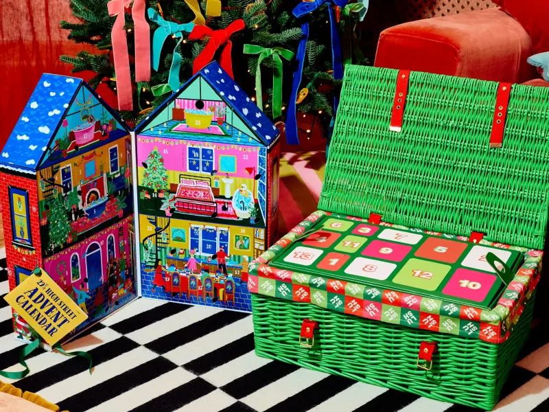 Colorful advent calendars designed by Lush, featuring a festive house and a green wicker basket, on a striped rug.