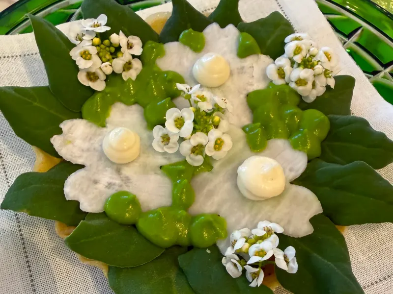 A beautifully arranged dessert featuring edible flowers, green leaf decorations, and creamy dollops on a cloth napkin.