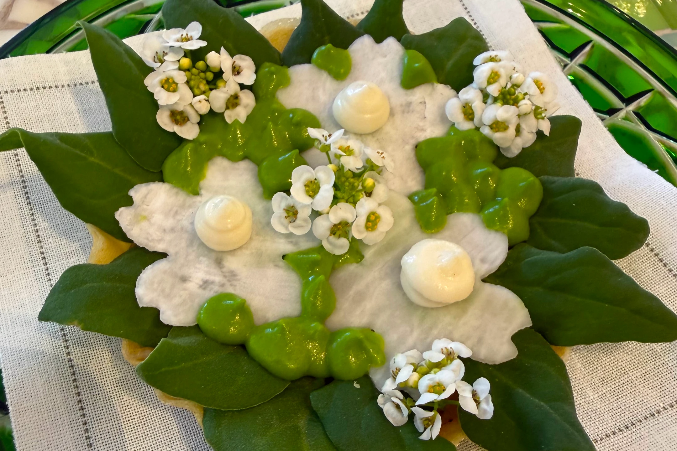 A beautifully arranged dessert featuring edible flowers, green leaf decorations, and creamy dollops on a cloth napkin.