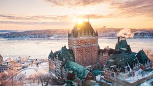 Fairmont Le Château Frontenac illuminated by sunrise, overlooking the frozen St. Lawrence River and cityscape in winter.