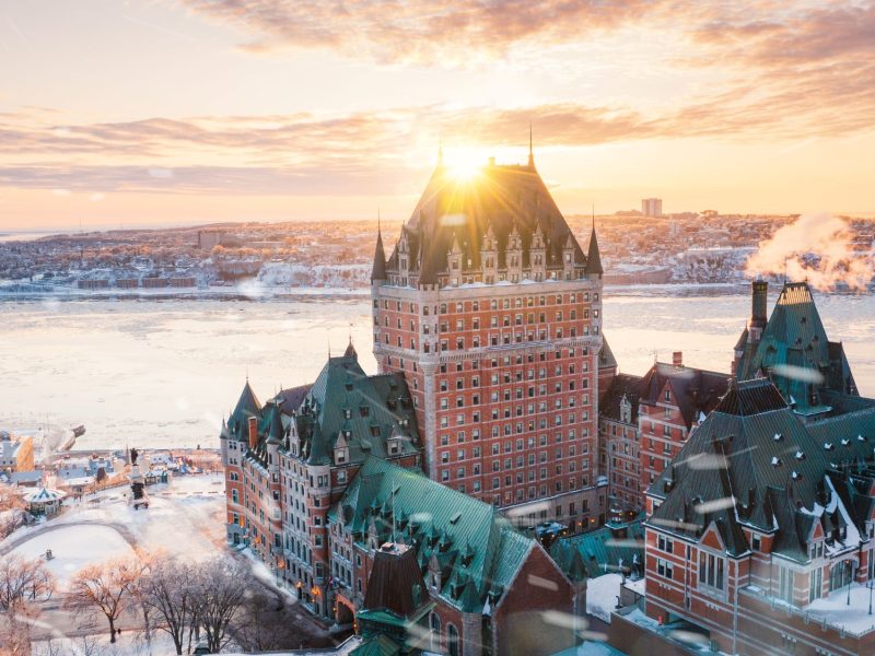 Fairmont Le Château Frontenac illuminated by sunrise, overlooking the frozen St. Lawrence River and cityscape in winter.