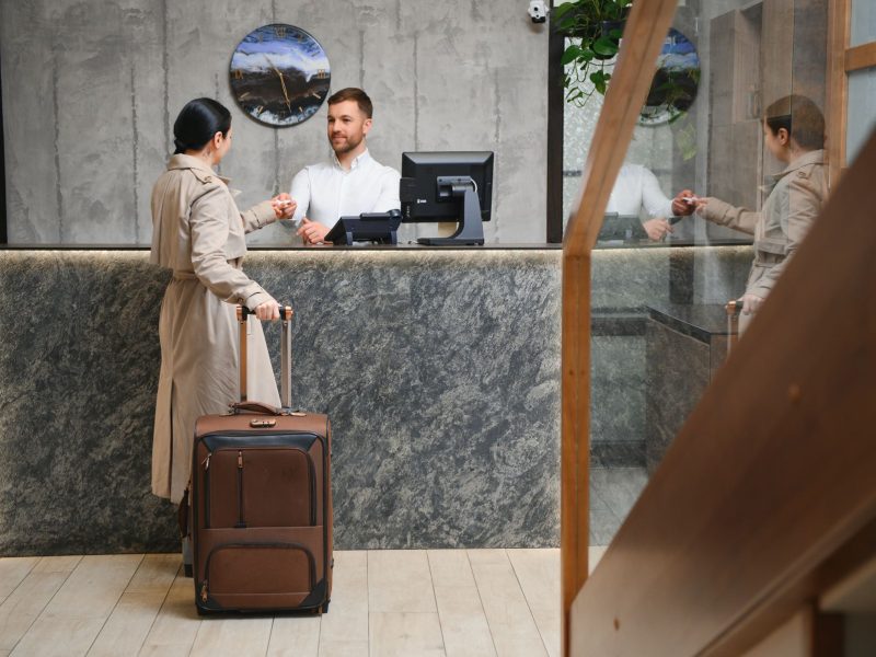 A traveler at a hotel reception desk hands a card to the clerk while holding a suitcase.