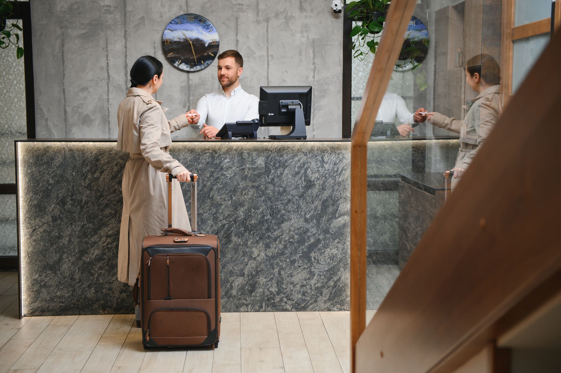 A traveler at a hotel reception desk hands a card to the clerk while holding a suitcase.