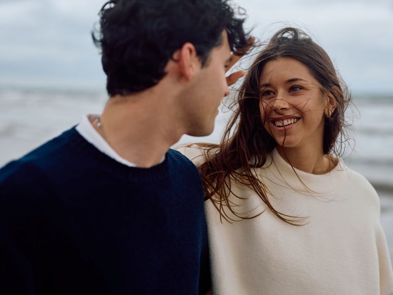 A couple shares a joyful moment on the beach, with wind-blown hair and smiles, illustrating connection and first impressions.