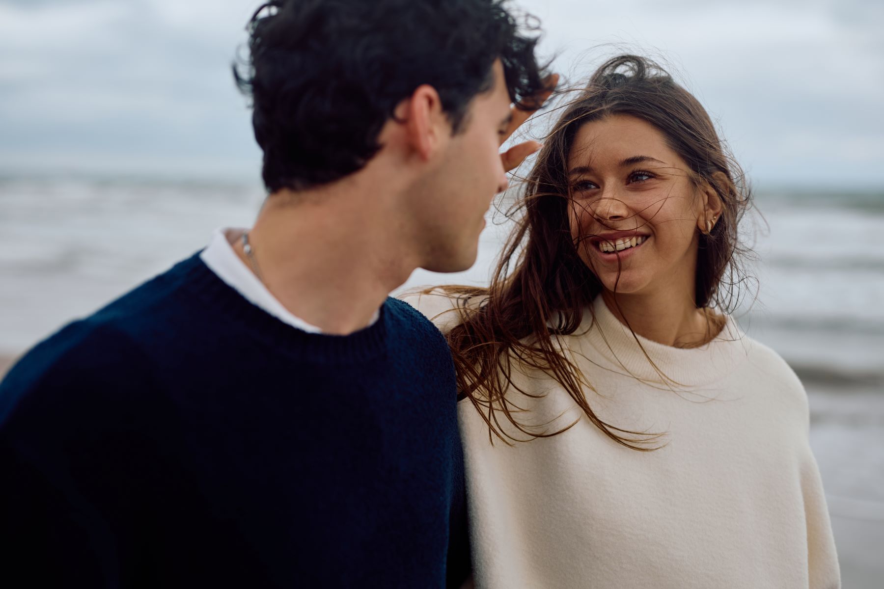 A couple shares a joyful moment on the beach, with wind-blown hair and smiles, illustrating connection and first impressions.