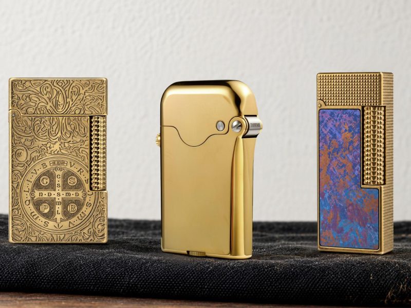 Three luxury lighters in gold and decorative designs displayed on a dark fabric surface.