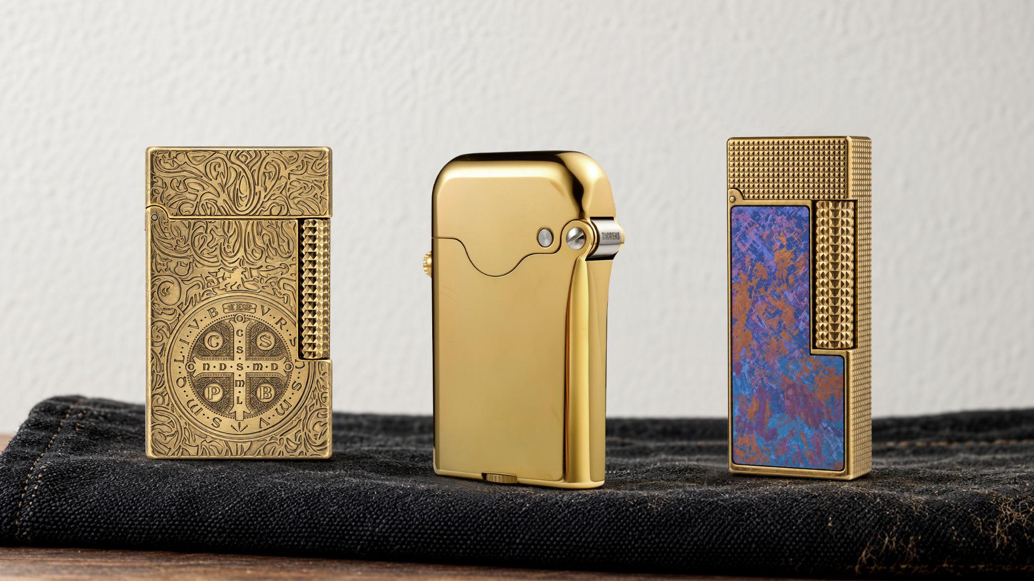 Three luxury lighters in gold and decorative designs displayed on a dark fabric surface.