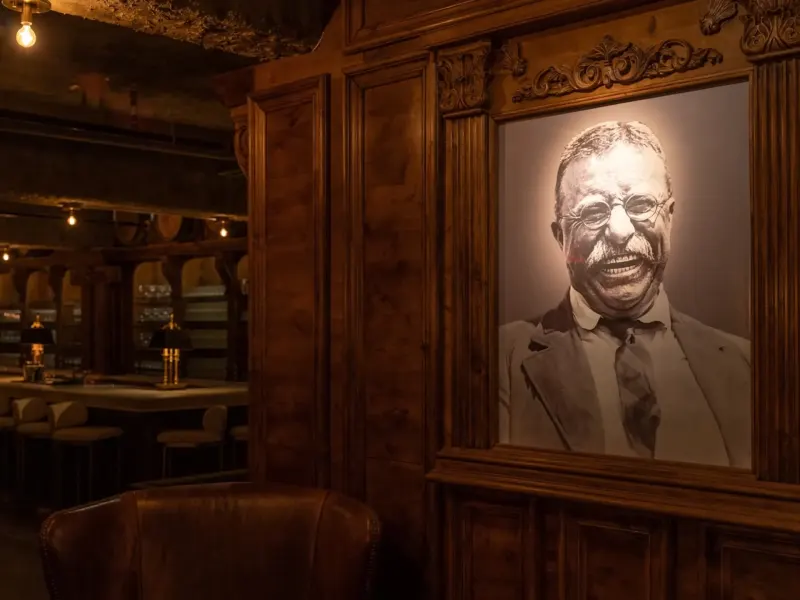 Framed portrait of Theodore Roosevelt in a cozy, wooden-panelled speakeasy setting with warm lighting.