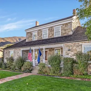Historic stone house with flags, a porch, and a landscaped yard, set against a backdrop of mountains.