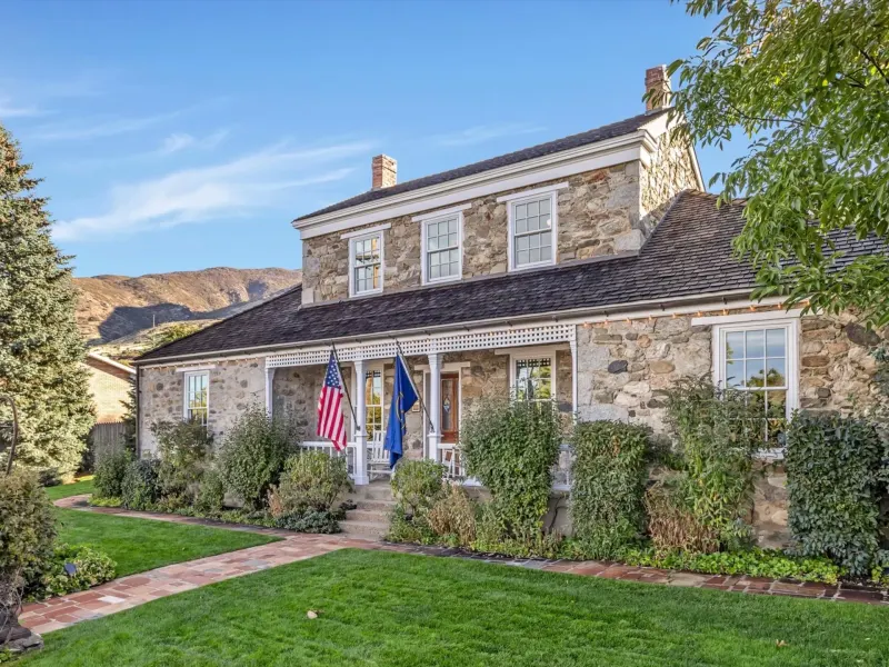 Historic stone house with flags, a porch, and a landscaped yard, set against a backdrop of mountains.