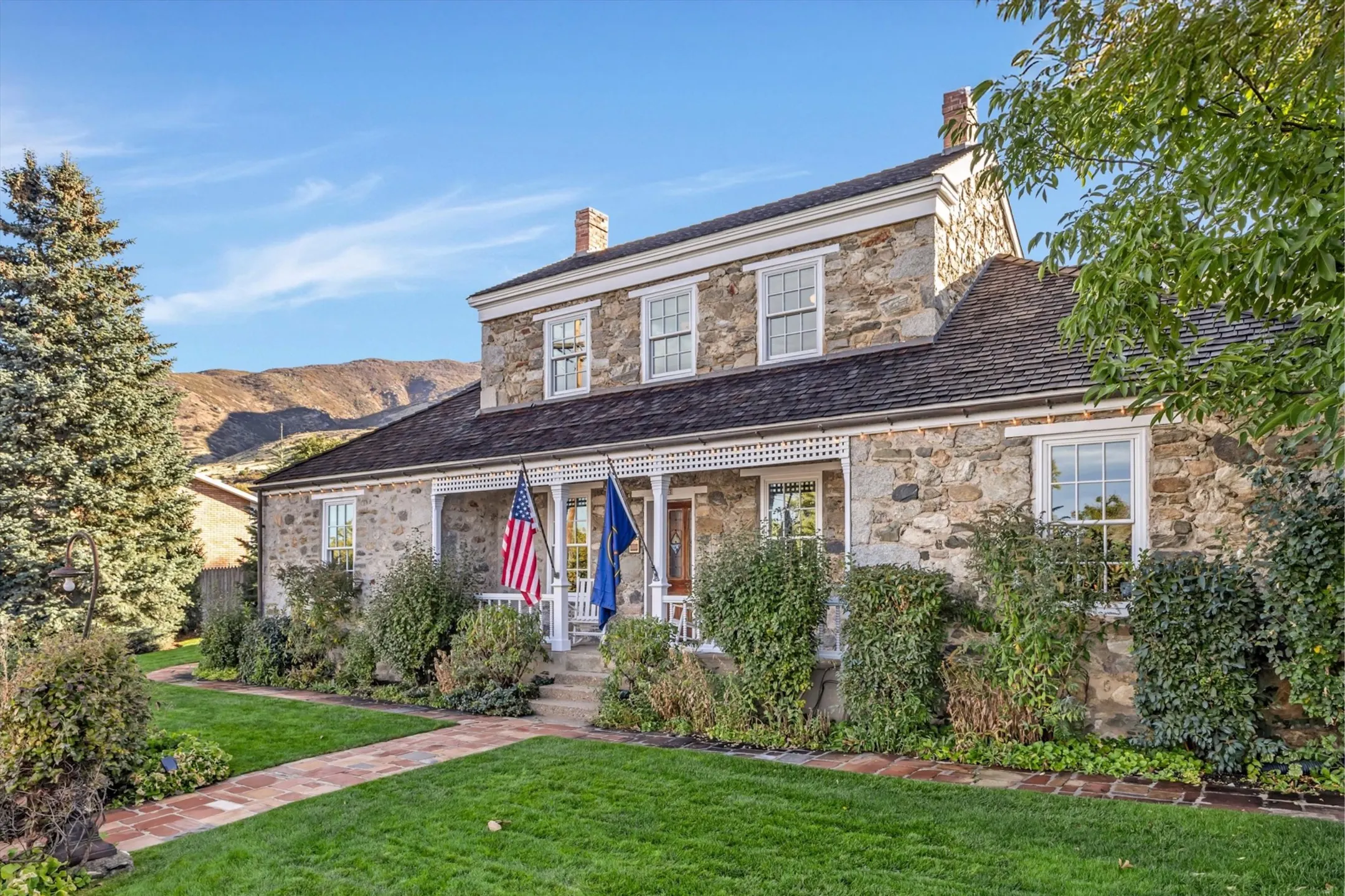 Historic stone house with flags, a porch, and a landscaped yard, set against a backdrop of mountains.