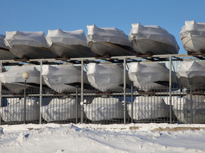 Boats wrapped in protective covers stacked on racks, preparing for winter storage in a snowy environment.
