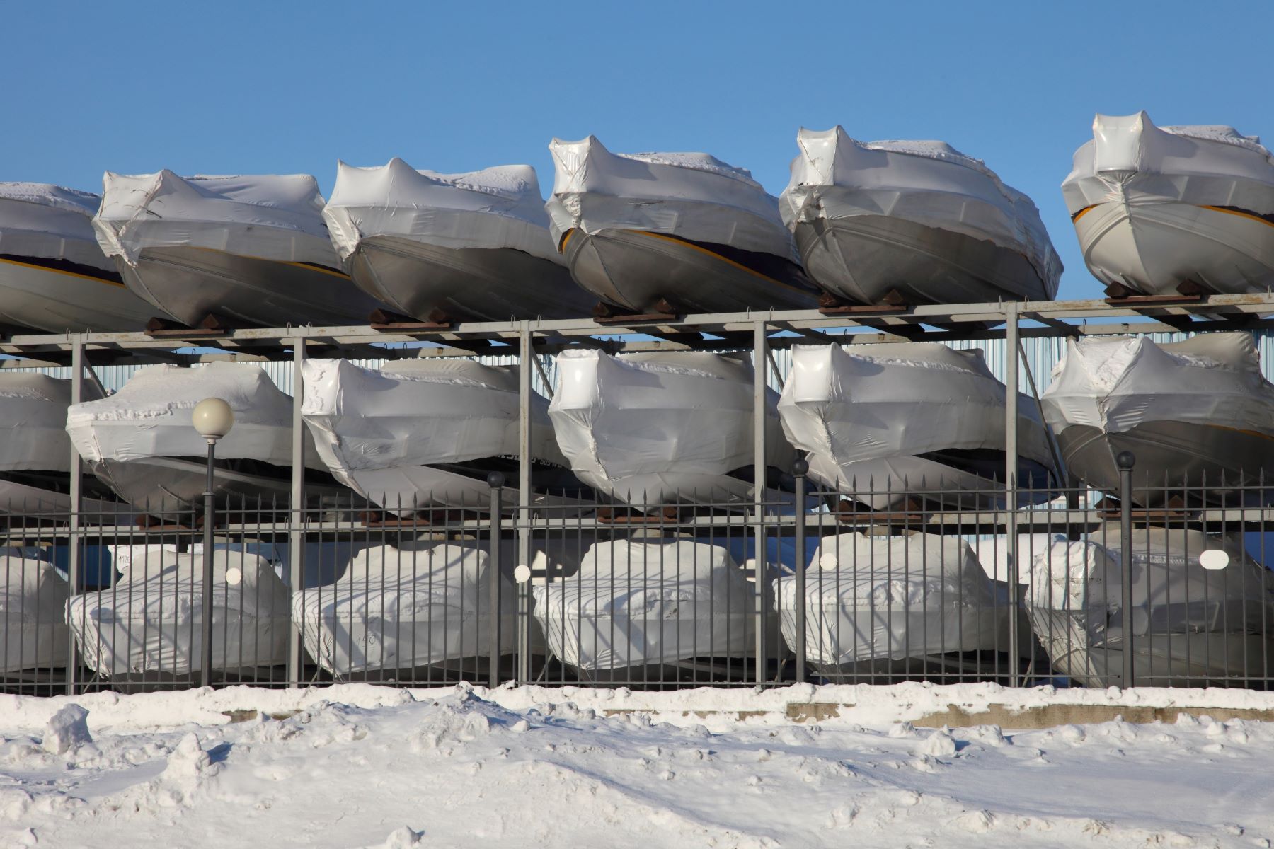 Boats wrapped in protective covers stacked on racks, preparing for winter storage in a snowy environment.