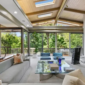 Bright, modern living room featuring large windows, a glass table, and cozy seating, overlooking lush greenery outside.