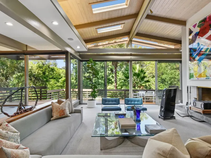 Bright, modern living room featuring large windows, a glass table, and cozy seating, overlooking lush greenery outside.