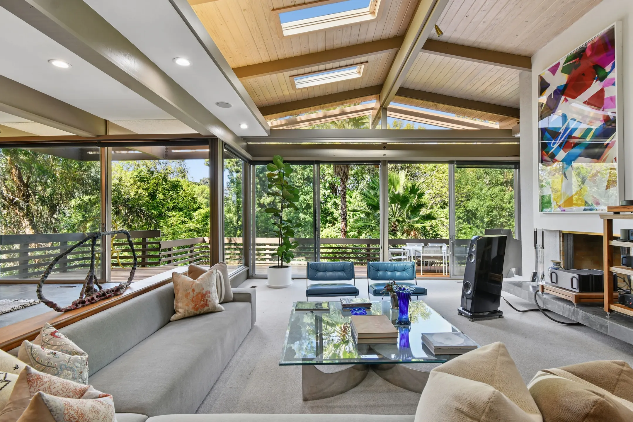 Bright, modern living room featuring large windows, a glass table, and cozy seating, overlooking lush greenery outside.