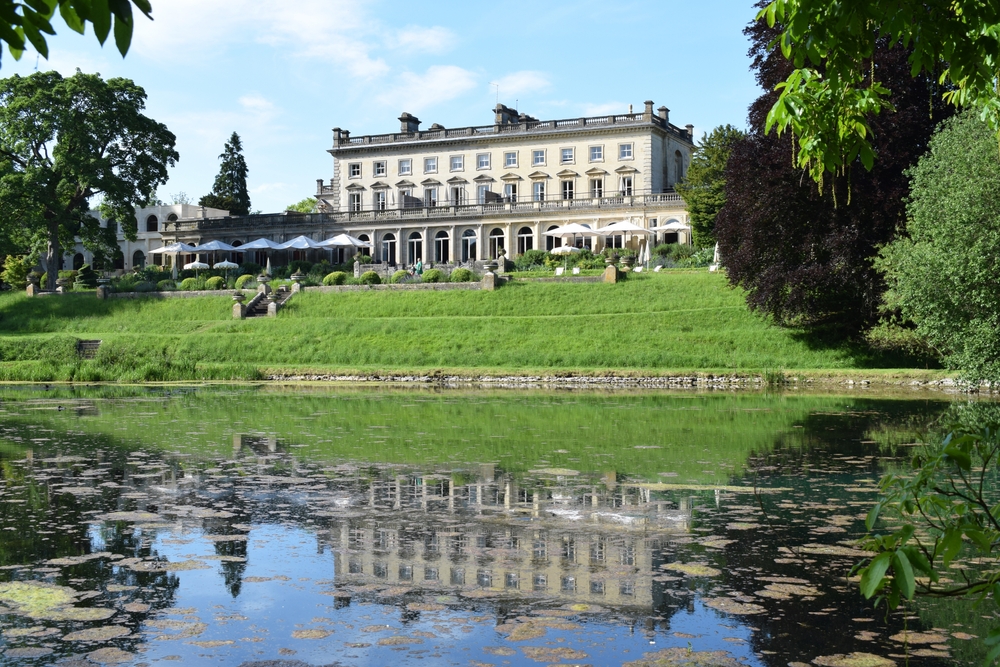 Historic hotel reflecting in a tranquil pond, surrounded by lush greenery and outdoor seating for guests.